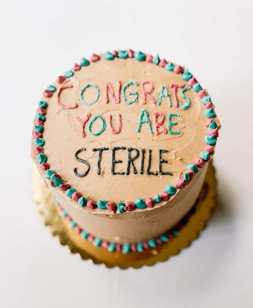 A light brown cake with pink and blue dots on the top and bottom edges. On top it says "congrats you are" in alternating pink and blue and under it says "sterile" in black