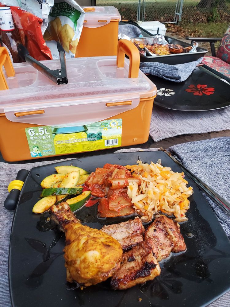 A plate on an outdoor table with the post mentioned foods on it of chicken drummies, pork belly, pickled veggies, kimchi, and cucumbers with chili oil. Other empty plates and pickling jars are on the table as well.