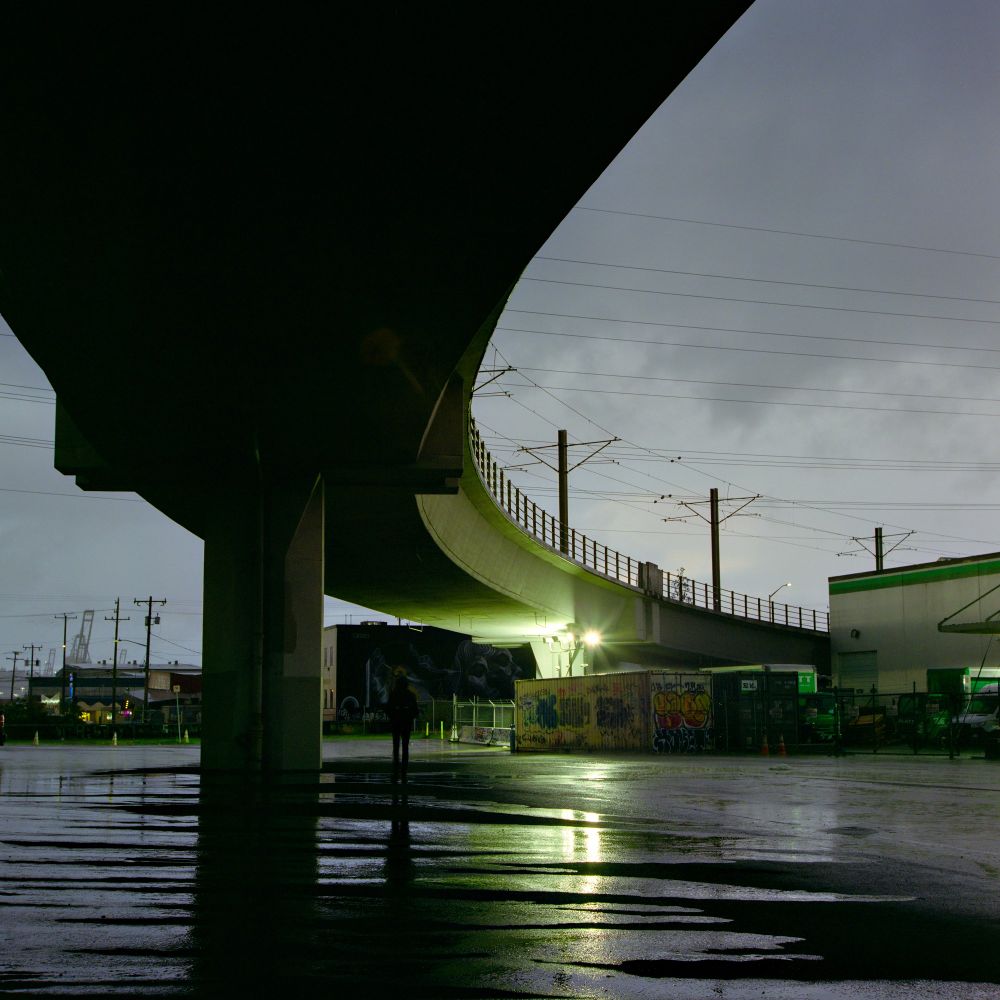 An anonymous figure hides from the rain under an overpass