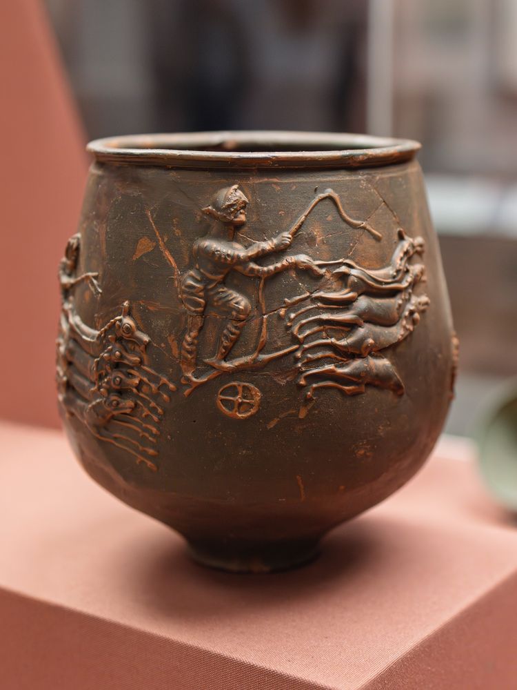 Large cup or jar, dark brown (it would have been glossy black when new) depicting a chariot race in barbotine decoration, which unsurprisingly looks a lot like cake icing details, because it's piped onto the cup in the same way, with a bag. The auriga - charioteer - wears long-sleeved jerkin and leggings, and a leather helmet. He holds a whip in his right hand, and the reins of the four horses in his left. Another chariot is fast on his heels. 

Found at Colchester, Essex. British Museum (1857,0806.1)
