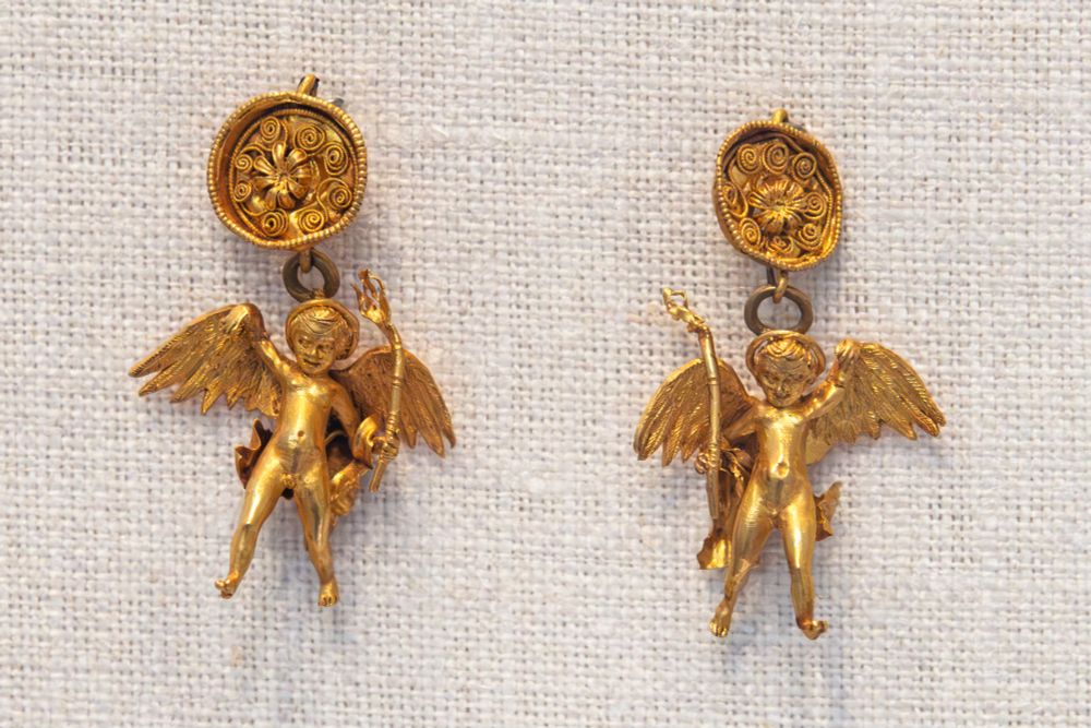 A pair of gold earrings, part of the 'Bastis Gold' treasure. Each earring has a floral disk - on the back of which is a hook - from which an Erote is suspended. The laughing winged Erotes carry torches, perhaps symbolizing ardent desire and burning love. Tremendous details in the face, fingers and toes - the Erotes also mirror one another.

Greek, Hellenistic, late 4th-3rd century BCE.

width of a: 3/4 in. (1.9 cm)
width of b: 15/16 in. (2.4 cm)

Met Museum, New York  (1995.539.8a, b)