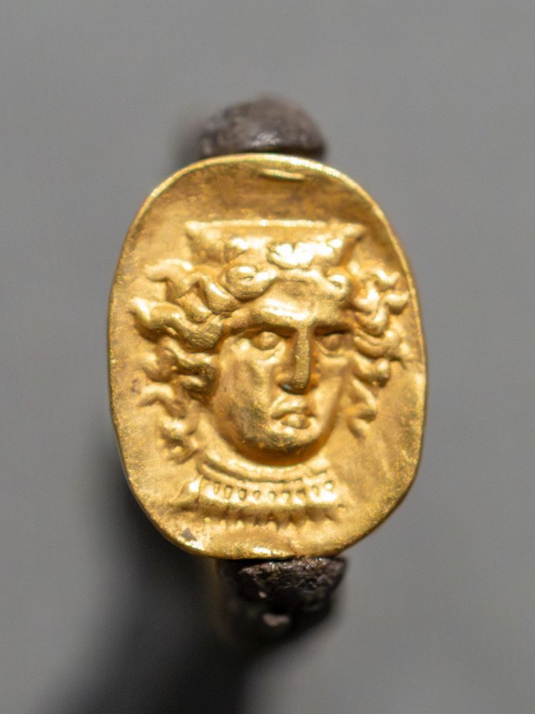 The head of a goddess decorates the flat surface of this hollow gold scarab set in a silver swivel ring. Carved in relief rather than engraved as on a stone scarab, the goddess wears a stephane or crown decorated with three palmettes and a necklace with acorn-shaped pendants. Her locks of wavy hair radiate out from her face and she has a fierce expression. She represents Hera Lakinia, a local version of Hera, whose sanctuary was near the city of Kroton in South Italy. Coins of Kroton from the 300s BCE show similar images of Hera Lakinia, supporting this identification. 

Getty Villa Museum (85.AM.273)