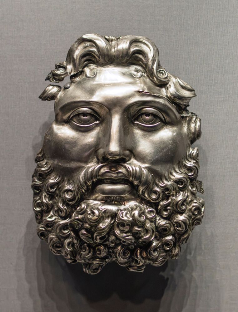 Mask-like portrayal of a mature man with long wavy hair parted at the center, and a luxuriant curling beard. The face is broad, with a straight nose and parted lips. The eyes are heavily-lidded, with the outline of the iris incised and shallow insets for the pupils. In many respects, the face resembles depictions of Jupiter (the Greek Zeus) as Serapis, an Egyptian god of fertility adopted by the Greeks in the Hellenistic period. The function of this relief is uncertain, but it may have been part of a large decorative element on a ceremonial shield or another piece of armor.

Roman, ca. 1-50 CE. Silver.

18 × 13 × 5 cm (7 1/16 × 5 1/8 × 1 15/16 in.)

Getty Villa Museum (82.AM.88)