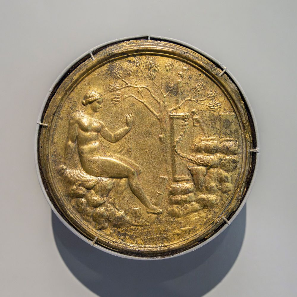 Artemis (the Roman Diana), or a Roman lady with divine fantasies, after her bath in a rustic, woodland setting, is the subject of the tondo in relief on the back of this Roman hand mirror. Her cloak is draped over the rocks on which she sits, and she holds the end wrapped around a small hand mirror in her raised left hand, a divine celebration of the uses of the mirror in a Roman household. The landscape in front of her, to the right, recalls the paintings and reliefs from houses around the Bay of Naples before the eruption of Mount Vesuvius in A.D. 79. The quiver of the goddess leans against the base of a garlanded altar with a small herm on top. A second terminal figure, Priapis, the god of gardens and fertility, tilts back while facing to the right on the ledge at the right. The bovine skull in the right foreground suggests the sacrifice after a successful hunt.

Art Institute of Chicago (ARTIC) 1985.1042a-b