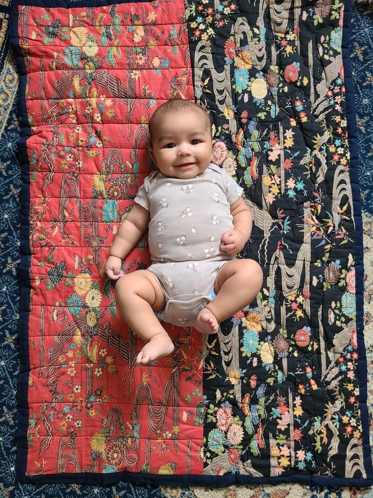 A smiling 3 month old baby lays on a quilted mat. The mat is sewn from Japanese printed quilting cotton and is hand quilted.