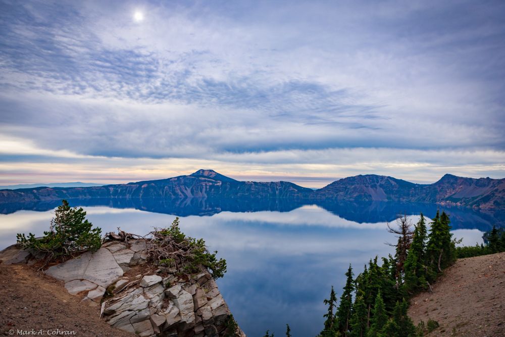 The still waters of Crater Lake reflect the cloudy sky in shades of blue and gray.