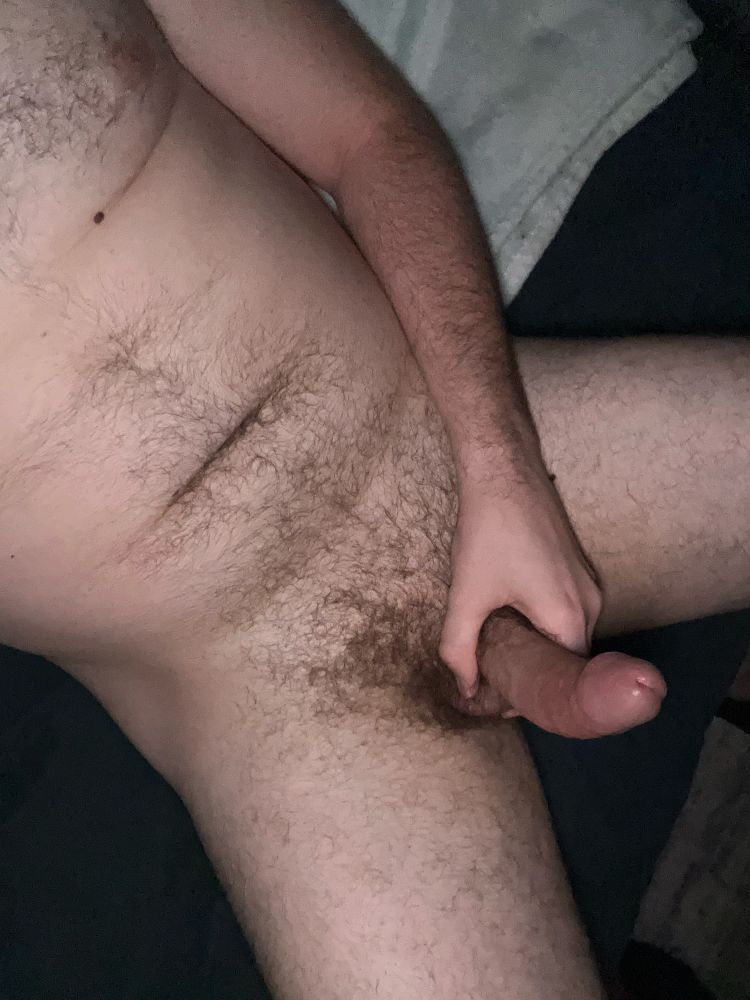 A view from above of a man sitting naked on a couch. The room is dark and the flash illuminates the man. He is moderately hairy and holding his erect cock with his left hand.