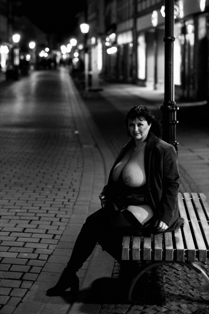Eine Frau auf einer Bank in einer nächtlichen Stadt. Die Brüste sind entblößt. Schwarzweiß. 