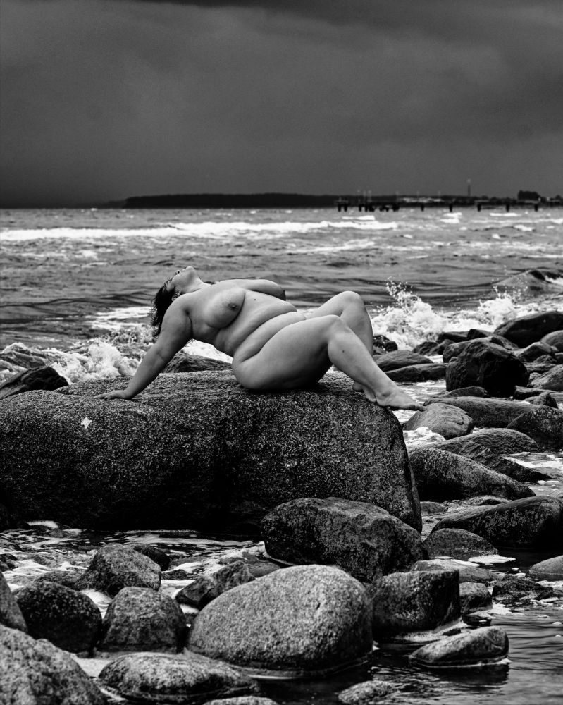 Eine nackte Frau liegt auf einem Felsen an der Ostsee vor einem dunklen Gewitterhimmel. Schwarzweiß. 