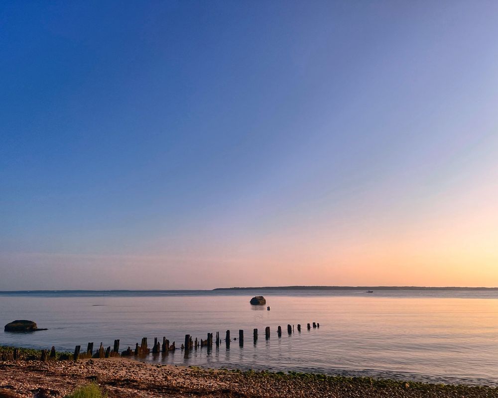 Gardiner’s Bay early morning. Calm water, old pilings jetting out from rocky beach. Pink glow to blue sky.