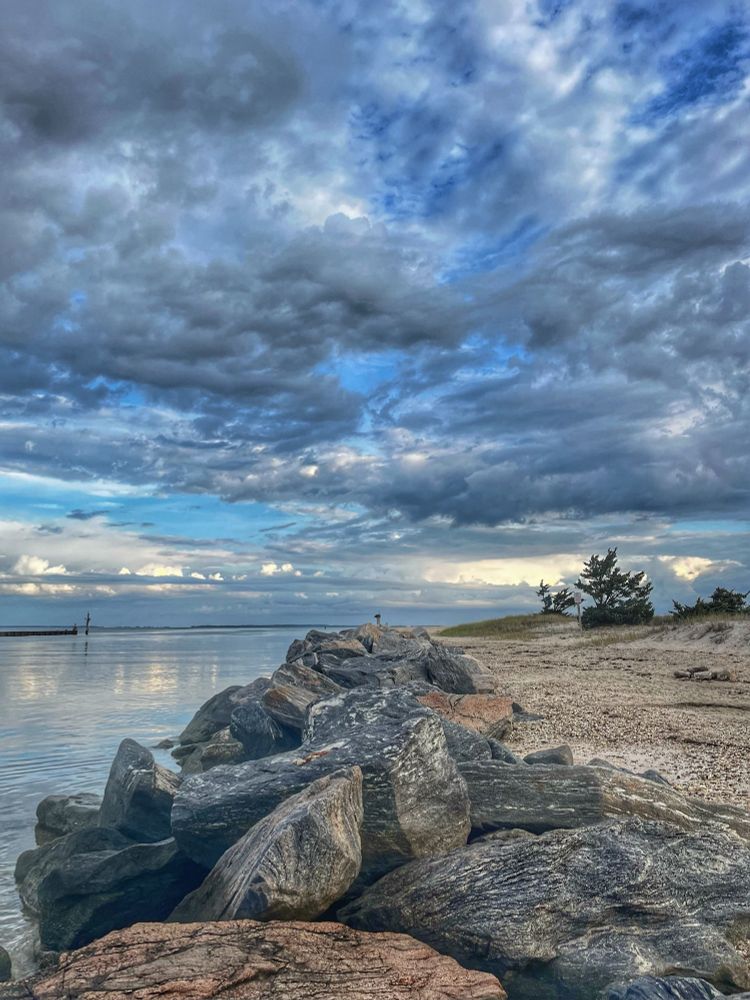 Rocky jetty, calm harbor water on left of jetty. Blue skies and clouds at end of jetty