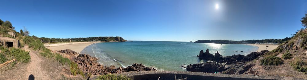 A panoramic view of St Brelades bay in Jersey (Channel Islands). The sun is shining bright at the top on the right side, causing a big white shimmer on the sea below. Surrounded are many trees, and 2 beaches on either side. Image taken from centre on iPhone 16.