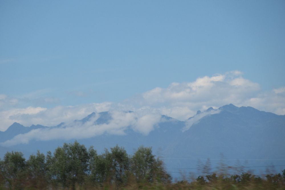 Photograph of a mountain range partially obscured by clouds, with trees in the foreground and a blue sky above.