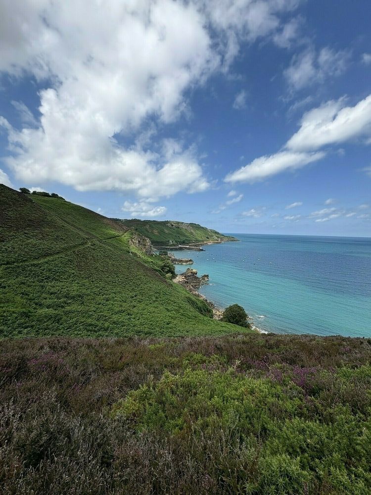 A scenic coastal landscape features a clear blue sea, rolling green hills, and a partly cloudy sky.