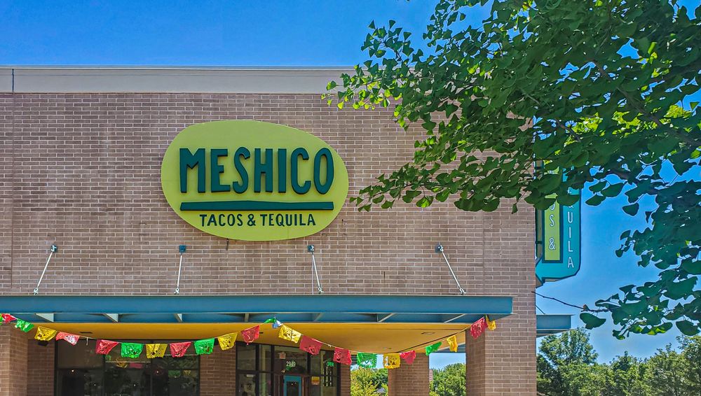 Oval-shaped sign with a light green background and dark green letters on the exterior facade of a brick-clad restaurant. Sign reads “Meshico Tacos & Tequila.”