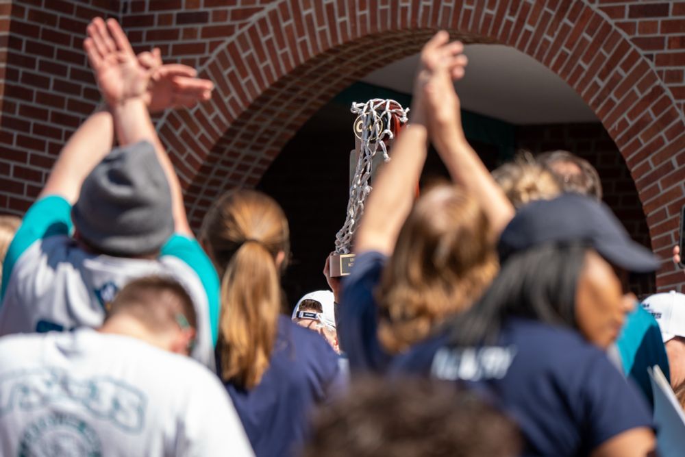 me the UNCW men’s basketball team back to the nest after winning the CAA Championship. The team arrives with the championship trophy, greeted by cheering fans. Players and coaches step off the bus, celebrating with the crowd as they prepare for their journey to the NCAA March Madness Tournament. Fans wave flags, hold signs, and capture the moment on their phones, filling the atmosphere with energy and school pride. 