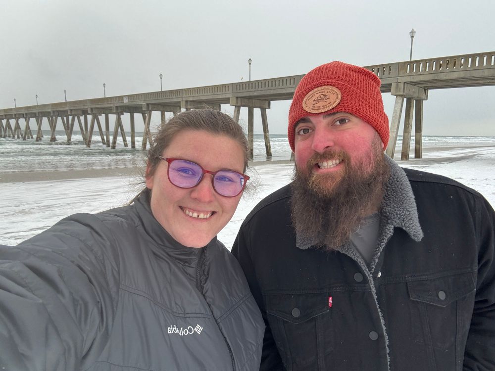 Selfie of two on a snowy beach in front of a concrete pier. 