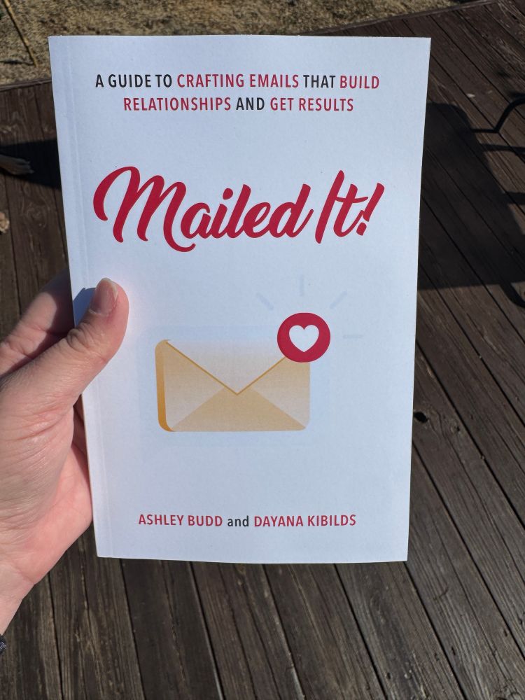 A person holding a book titled Mailed It! A Guide to Crafting Emails That Build Relationships and Get Results by Ashley Budd and Dayana Kibilds. The book has a white cover with a graphic of an envelope and a heart icon. The background features a wooden deck in natural light.