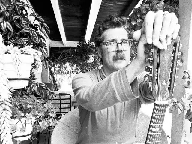 Black and white photo of Robbie Basho holding his guitar next to a bunch of potted plants