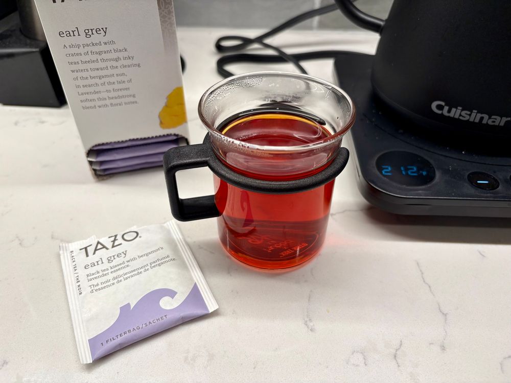 Tea, Earl Gray, Hot. A clear bistro glass with a black handle, as seen on Star Trek the next generation. It is sitting on a white marble countertop, next to an electric kettle, with a temperature gauge, indicating that the tea is hot. Next to the glass is an open packet indicating that the tea is Earl Gray.
