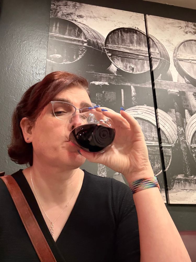 A picture of our poster, relaxing at a table, enjoying a glass of red wine, as is her divine right as a transbian wine aunt.