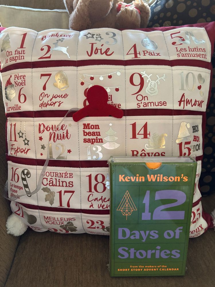 A green box containing Kevin Wilson’s 12 Days of Stories sits in front of a pillow advent calendar. The pillow is white with red lettering (in French) and with the silver and gold Christmas-y images.