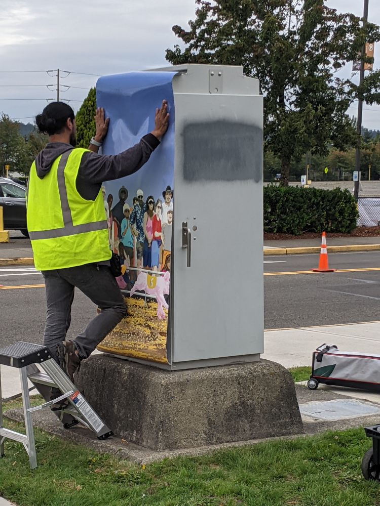 A worker installs the first vinyl sheet of a signal box wrap, which will depict a piglet race at the fair.  The art is at the same intersection as the state fair entry gate in Washington State.