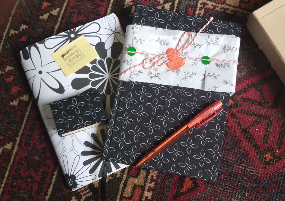 Dois cadernos já fora da caixa, uma agenda de capa preta com estampa floral abstrata branca e outro de capa de papel com motivo floral branco e preto, mais o bloquinho de notas adesivas e a caneta alaranjada