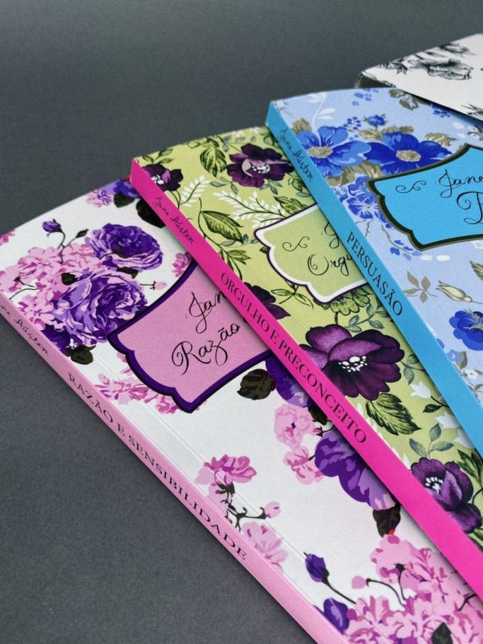 Imagem de três volumes de livros da Jane Austen, Orgulho e Preconceiro, Persuasão e Razão e Sensibilidade. Estão sobrepostos em leque, alinhado ao lado direito da imagem. As capas são padronizadas, com estampas de flores e ao centro um quadro com os títulos em fonte rebuscadas. As lombadas são de cor sólida, uma rosa claro, outra pink e outra azul.