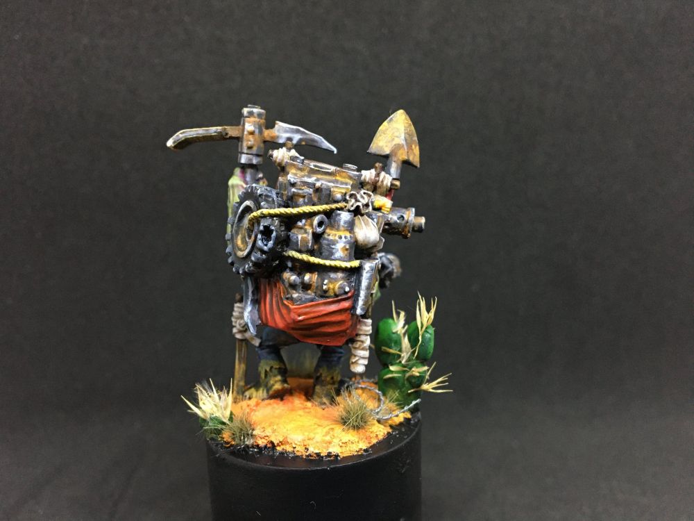 A painted miniature against a black background, depicting the overfull back-pack of the Orkish prospector. It is a red sack weighed down by rusty metal pieces.