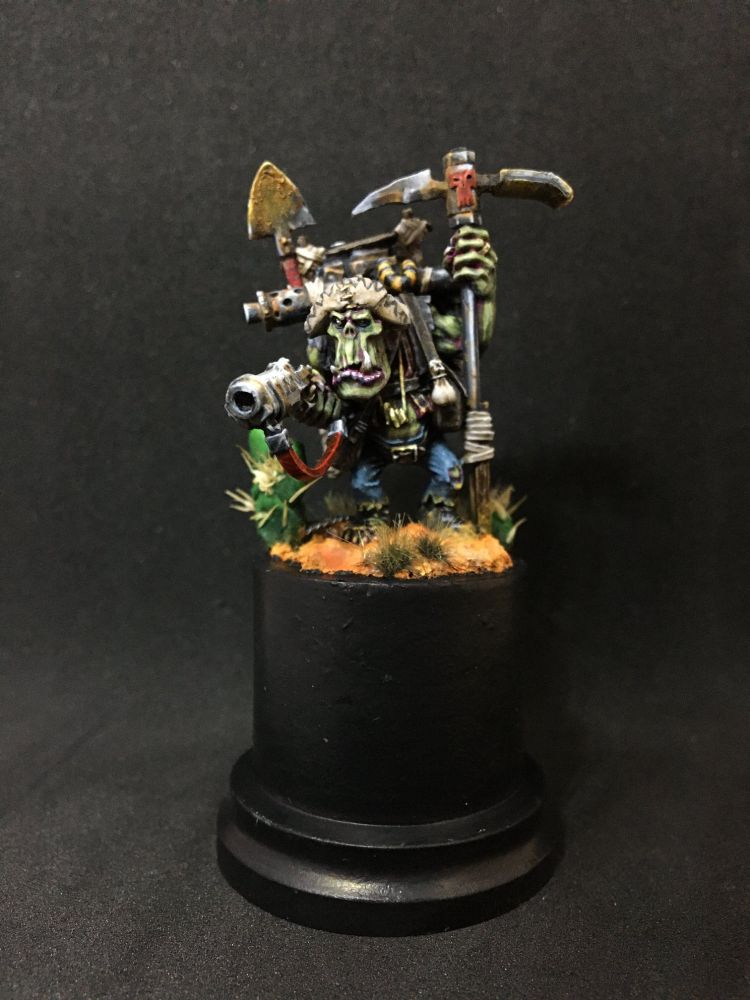 A painted miniature against a black background, depicting an Orkish prospector weilding a pickaxe and rusty gun. He stands on a orange desert base and wears a floppy sombrero.
