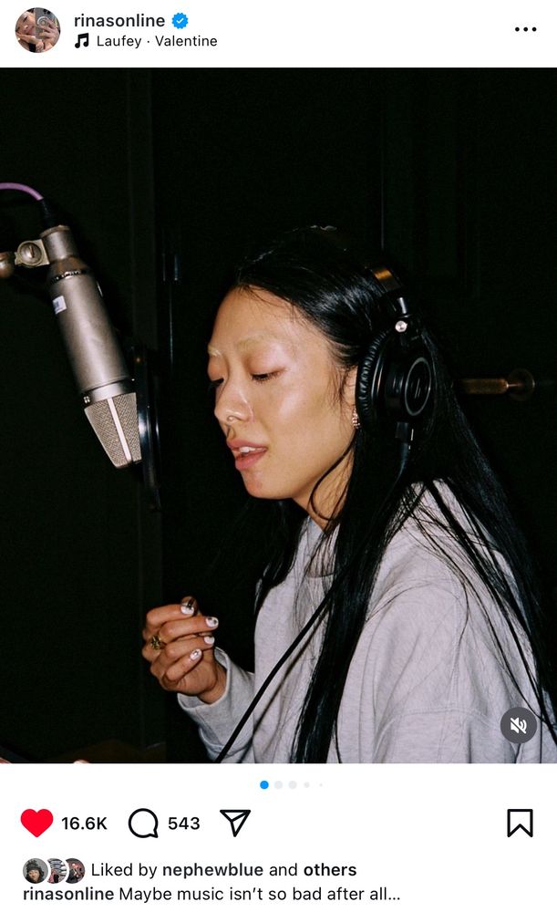 screenshot of Rina Sawayama’s Instagram post and she is in the studio with a mic close to her face and over-ear headphones on. She has long black hair and bleached eyebrows. 
The caption is “Maybe music isn’t so bad after all…”