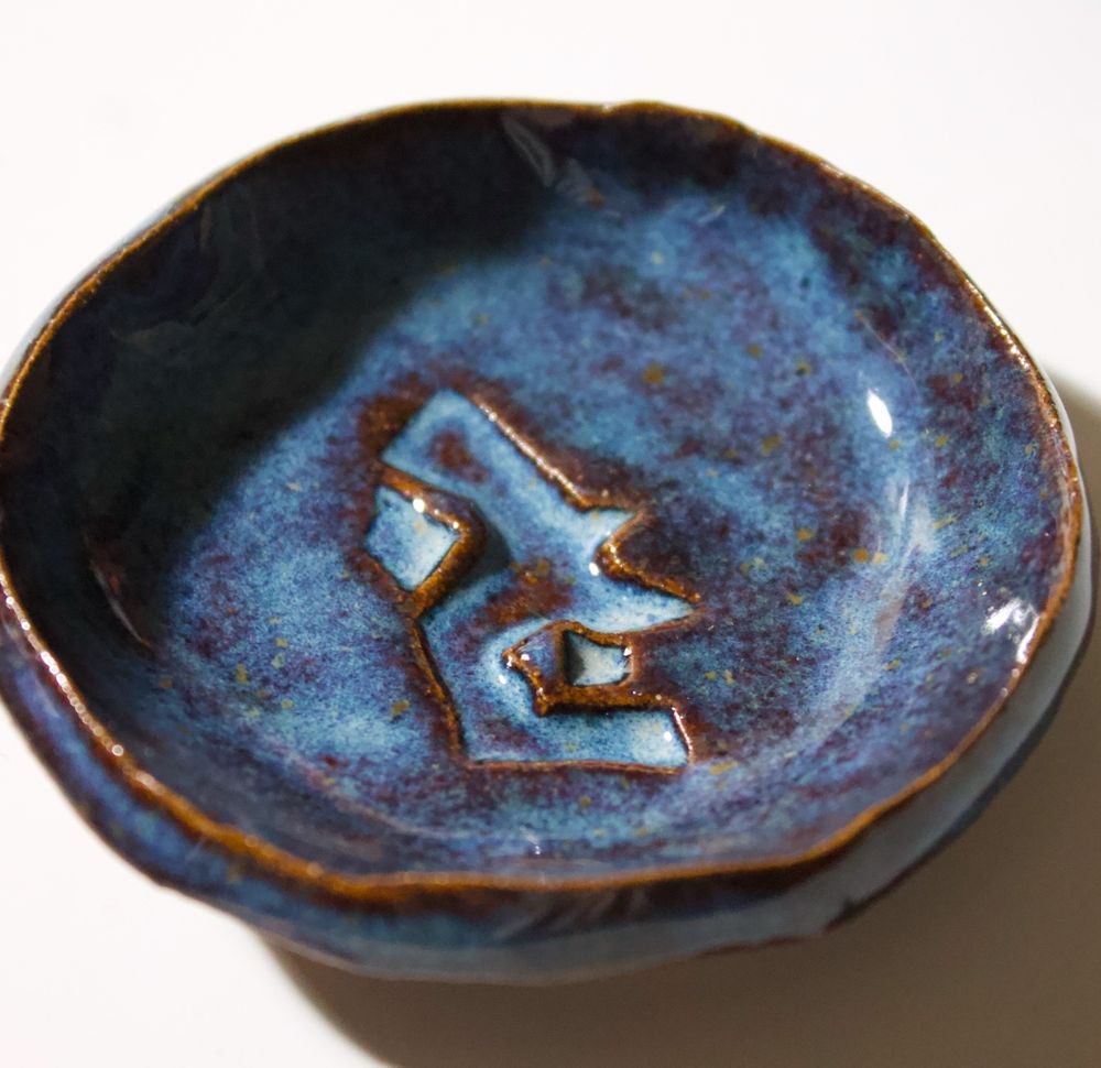 A finished ceramic dish with the Acceleration Rune from Arcane debossed into the surface. The glaze is a shiny deep blue-ish purple, with little yellowish flecks and a burnt effect along the edges. 