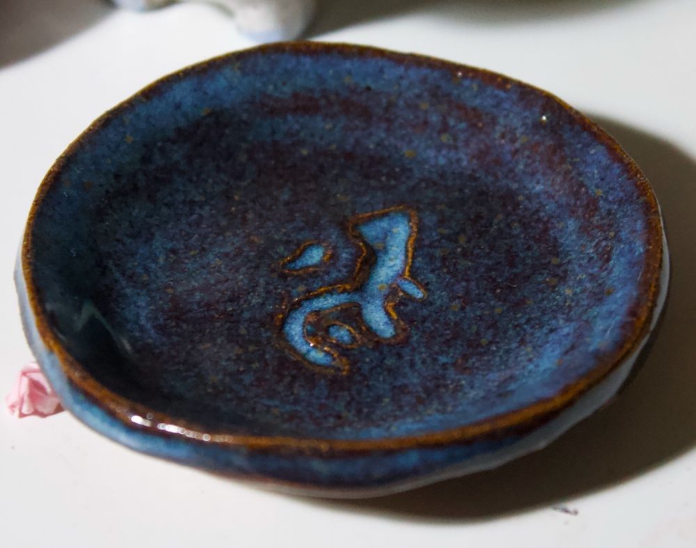 A finished ceramic dish with the Acceleration Rune from Arcane hand-carved into the surface. The glaze is a shiny deep blue-ish purple, with little yellowish flecks and a burnt effect along the edges. 
