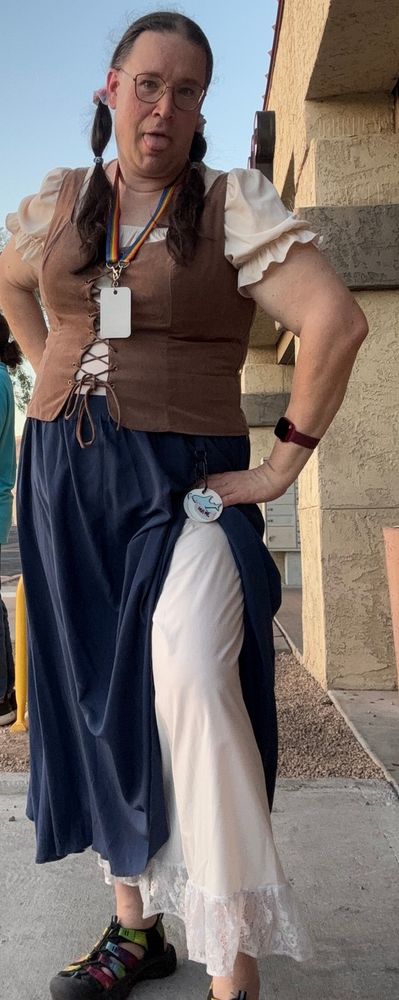 J.C. is wearing a renaissance themed outfit, with a two-layered skirt, a puff sleeve square top under a waistcoat vest, with pigtails on either side. She has the outer layer of the skirt hiked up on her left side to show off the petticoat or slip underneath it. She is also making a face at the camera because it's hot out and the camera and remote has been acting up on the phone and her smart watch. 