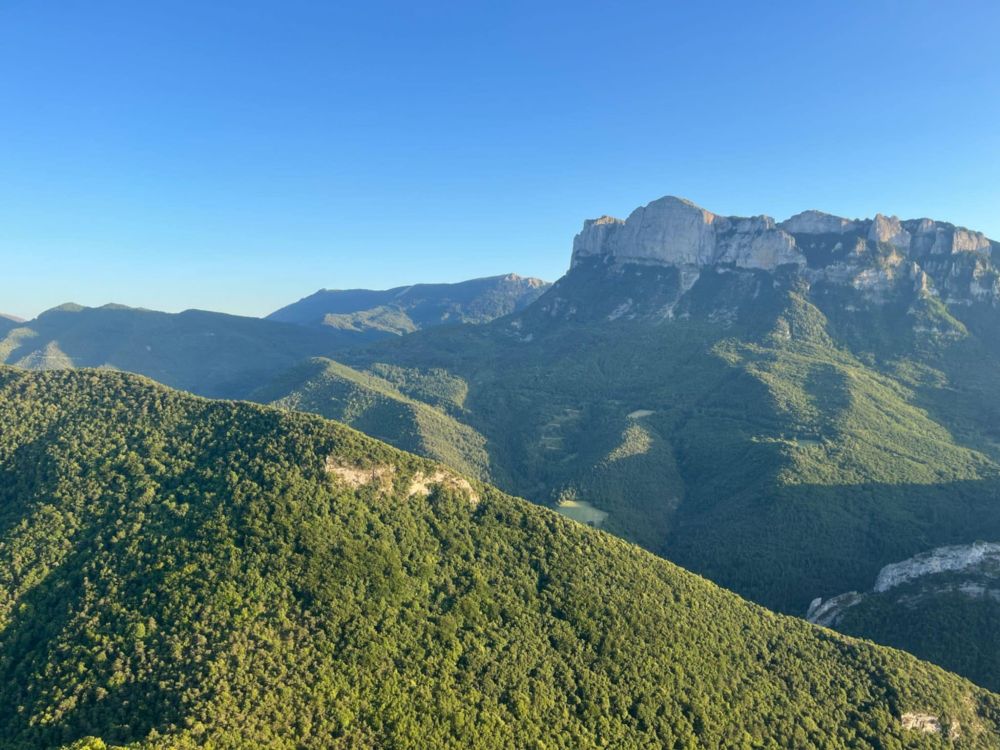 A view on top of a green mountain, with other mountains in the background. The sky is a clear blue