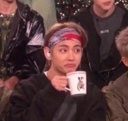 a blurry photo of V from BTS, a Korean man with light brown hair held back by a bandana. he is drinking out of a mug and staring off camera with a pinched expression that seems like he has a lot of opinions but won't share them 