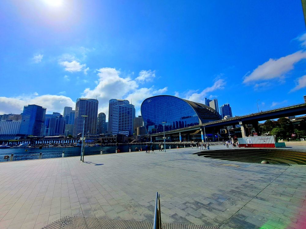 A view of big buildings, blue sky, white clouds, water, and pavement leading up to the harbor 