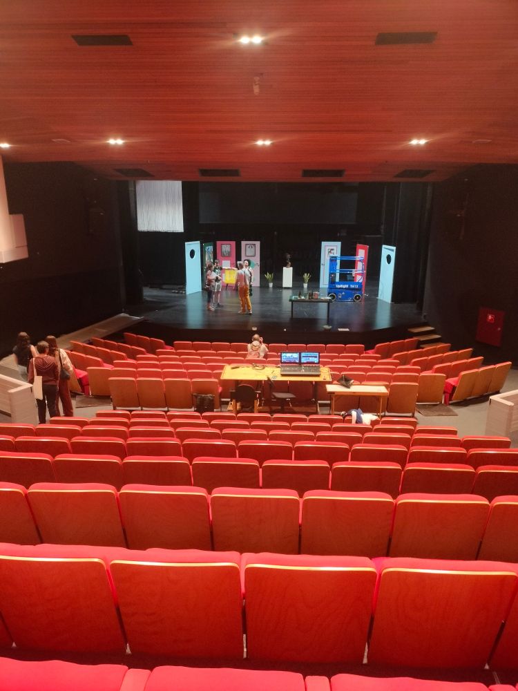 View of a stage from the back of the room, with empty red theatre chairs filling the room.