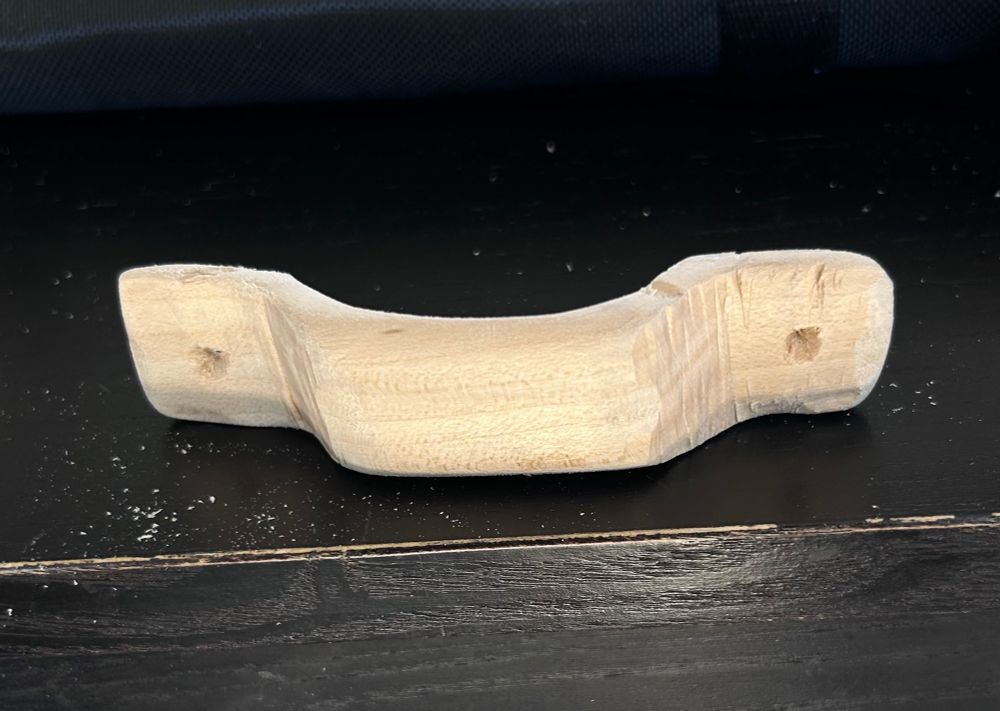 This is a close-up picture of a hand carved handle made from bassword. It sits on a black bench pilot holes have been started on the flat parts of the handle.