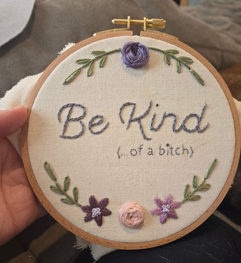 A piece of embroidery with flowers and leaves along the perimeter and text in the centre that says “be kind … of a bitch.”