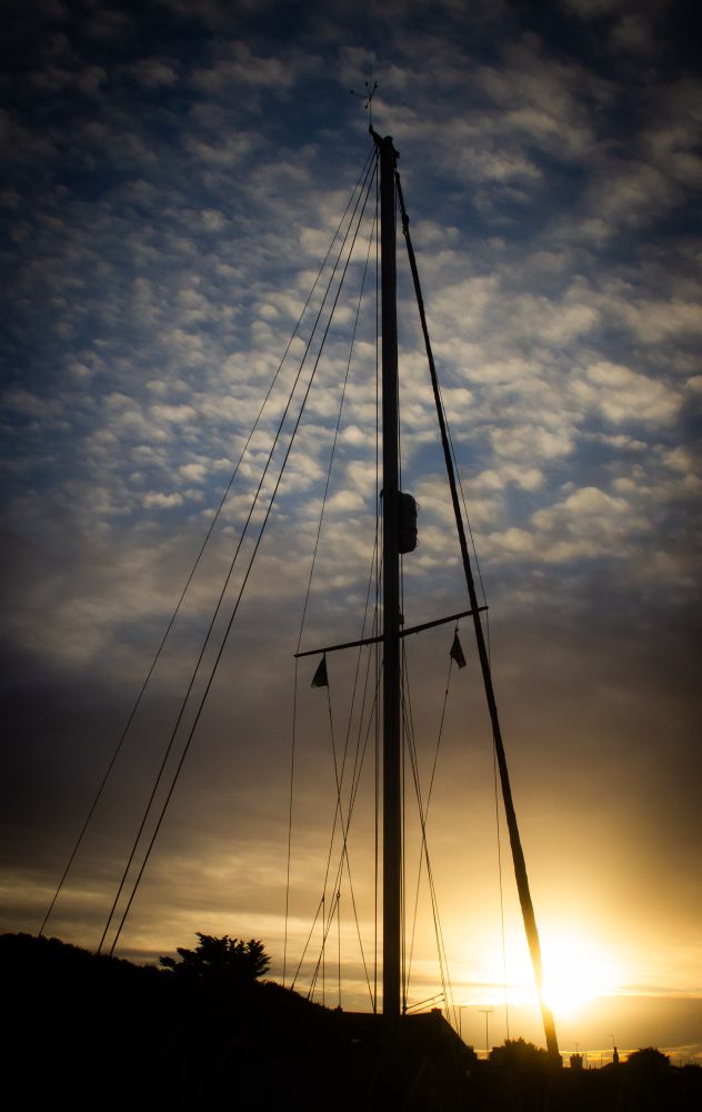 Silhouette of sailing boat mast stands tall in front of golden sunset in puff clouds filled sky. 