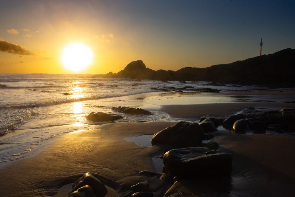 Sun flares across sky captured on Canon DSLR. Breakwater rocky structure with Barrel on pole tells all where Barrel Rock is. Flat sands bathe in golden sunlight and shallow tide. 