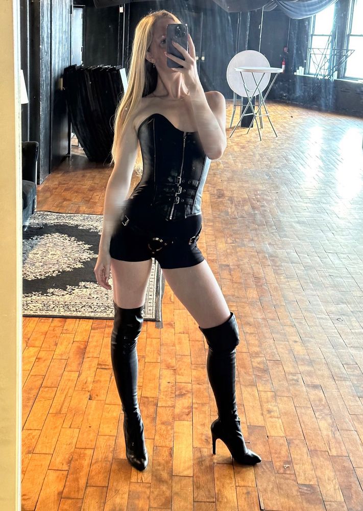 Princess Inés stands in front of a mirror with one leg cocked to the side and Her phone in front of Her face. She wears Her favorite thigh high leather boots, a new leather corset, tight black shorts and a leather harness. Her hair is straight.