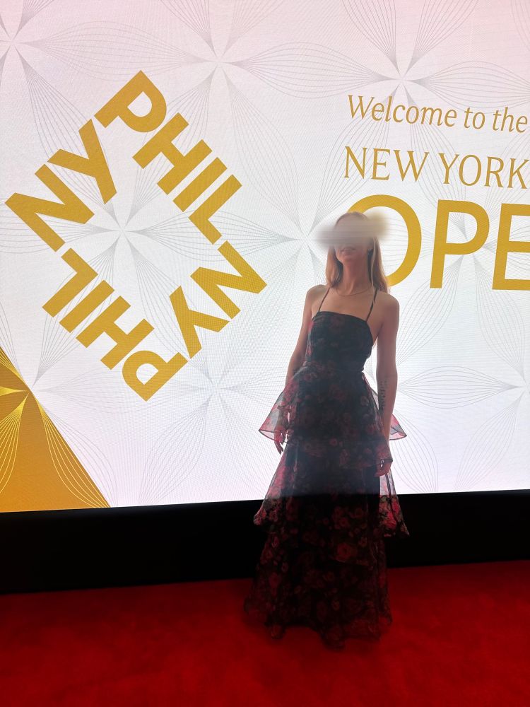Mistress Inés stands in a flowing, multi-layered red and black flowered gown on a red carpet in front of a NY Phil sign.