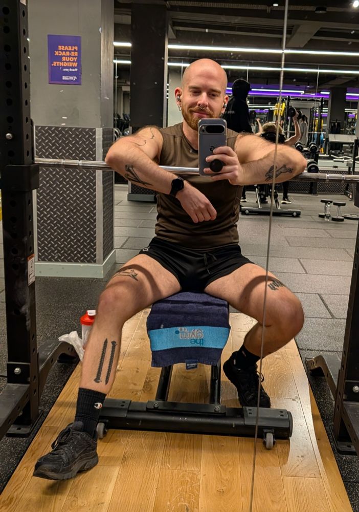 Man in shorts and tank top sat on the bench at the gym with his arms over the barbell.