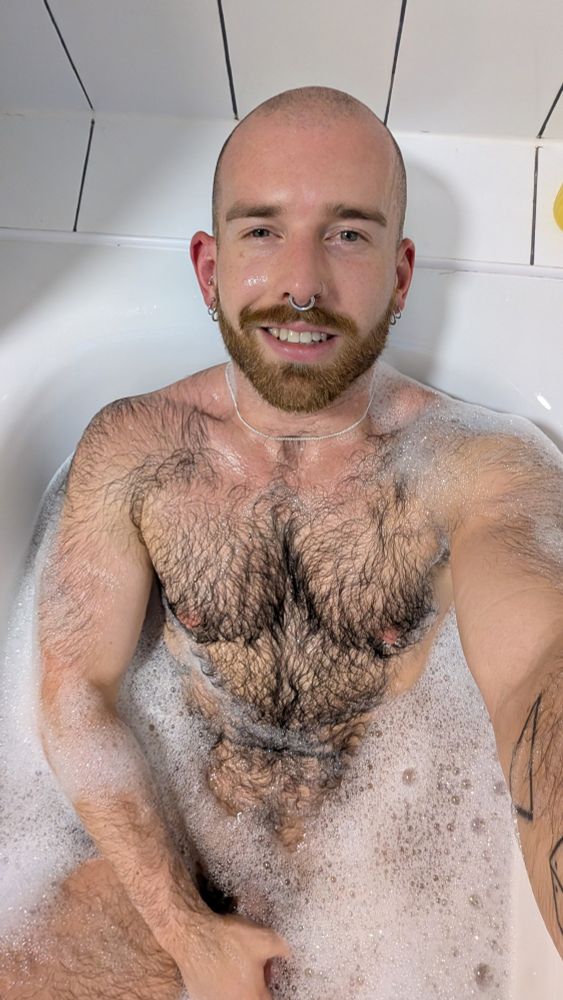A photo of a hairy bald man  with a ginger beard sat in a bubble bath 