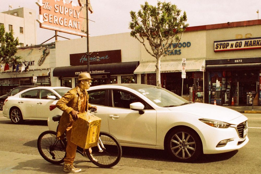A color photograph of a man body painted gold with matching gold clothing riding a bike on the street next to a white car