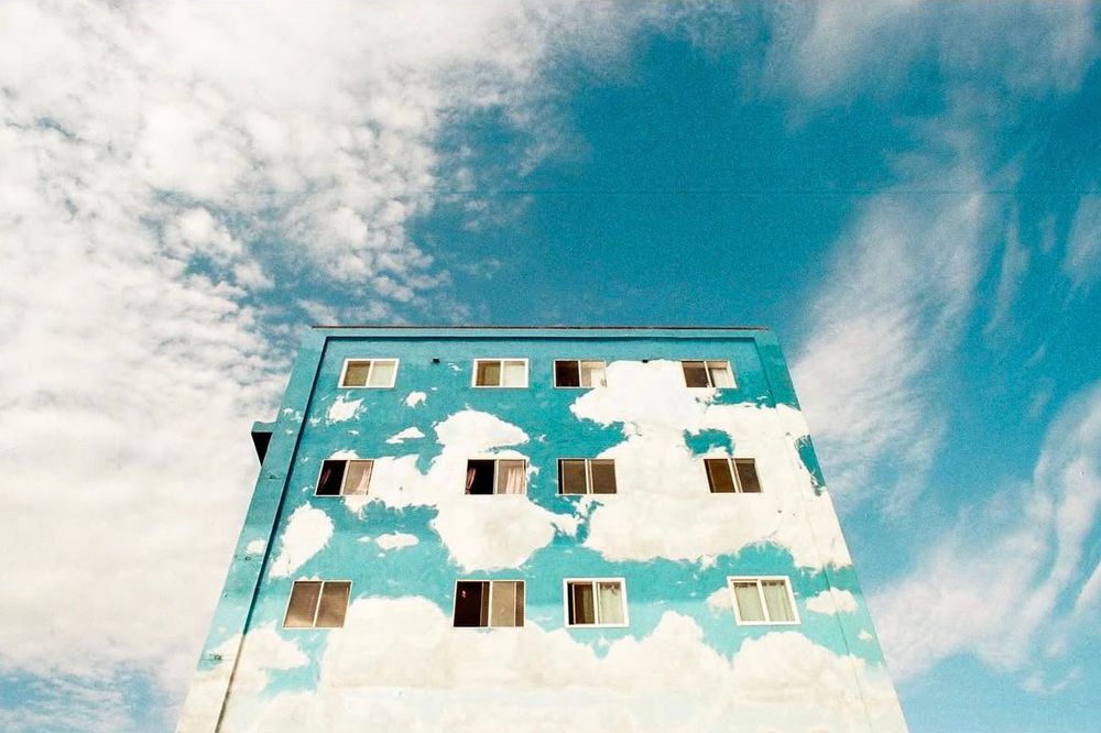A color photo of an apartment building painted as the sky with clouds while set against and contrasted by the natural sky with clouds