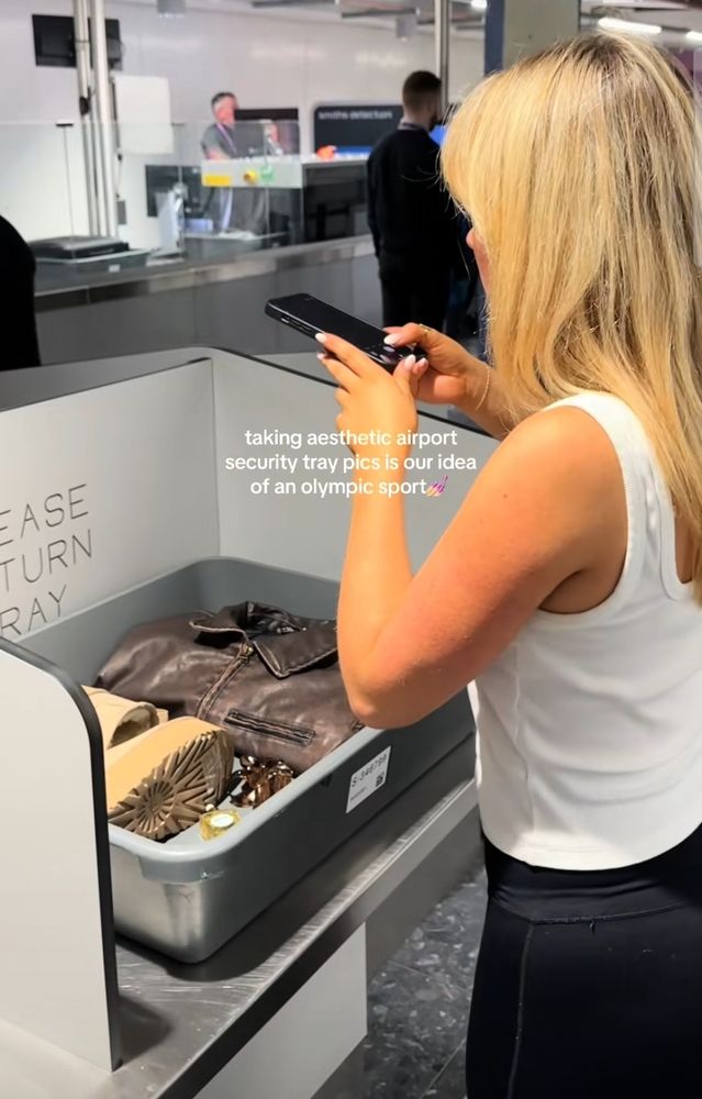 Picture of someone taking a picture of their airport security tray with the caption "taking aesthetic airport security tray pics is our idea of an Olympic sport 💅" 